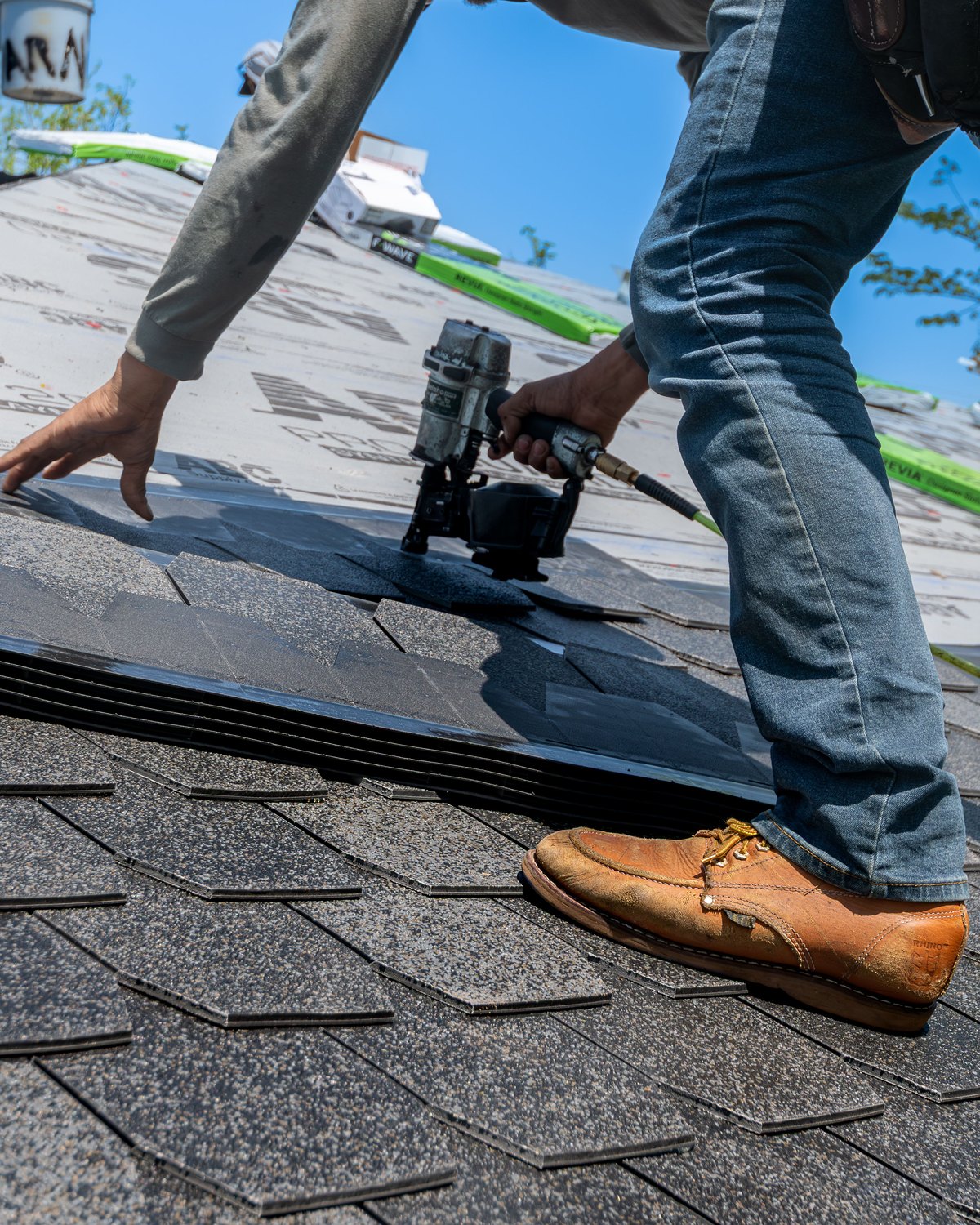 A roofer driving nails with a pneumatic nailer as he lays architectural shingles on a roof, under a clear sky.